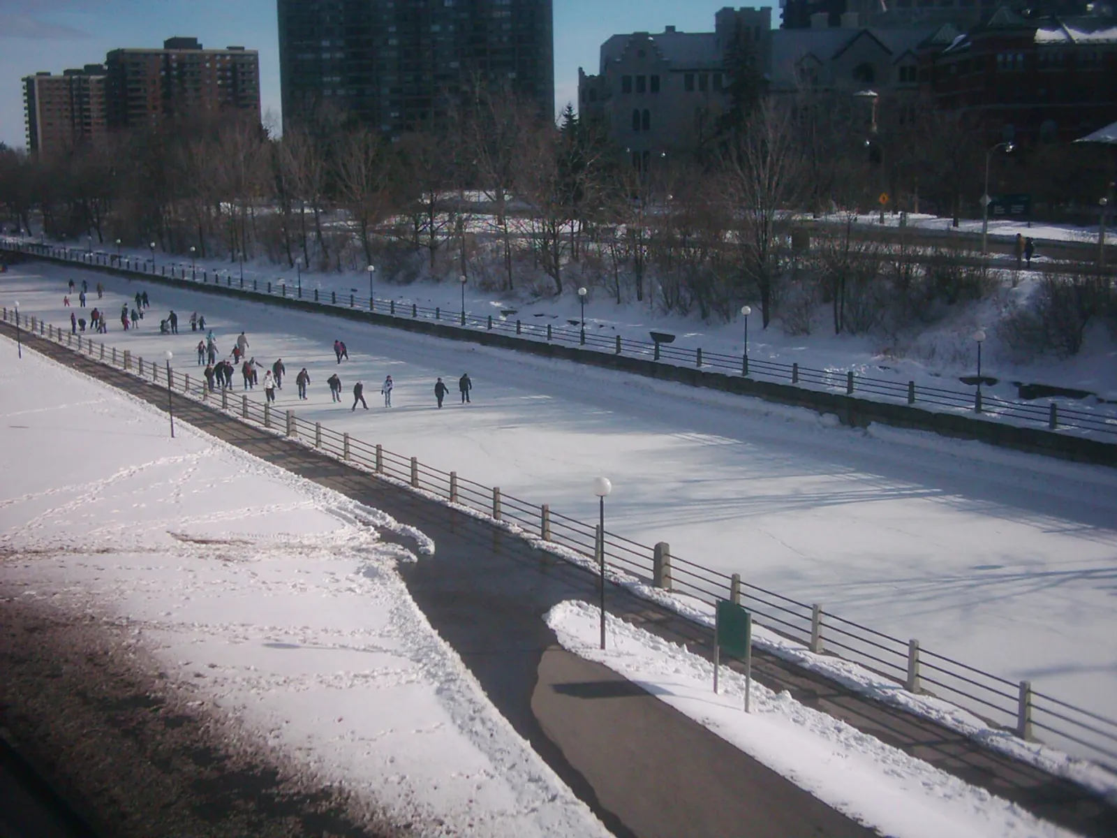 Vista a lo largo del Rideau Canal Skateway en invierno: un largo carril de hielo despejado junto a un camino nevado, con algunos patinadores y edificios de la ciudad más allá — Rideau Canal Skateway