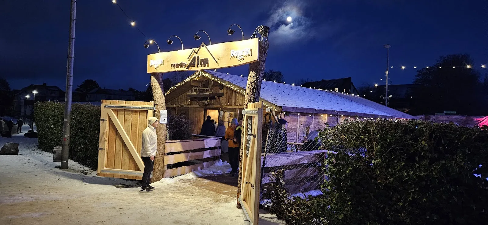 Das hölzerne Eingangstor und die Hütte der Eisbahn an einem mondhellen Abend, mit einer leichten Schneedecke auf den Dächern und einer Hecke neben dem Weg. – RIEDS Winterwelt Echteis-Eisbahn Roßdorf