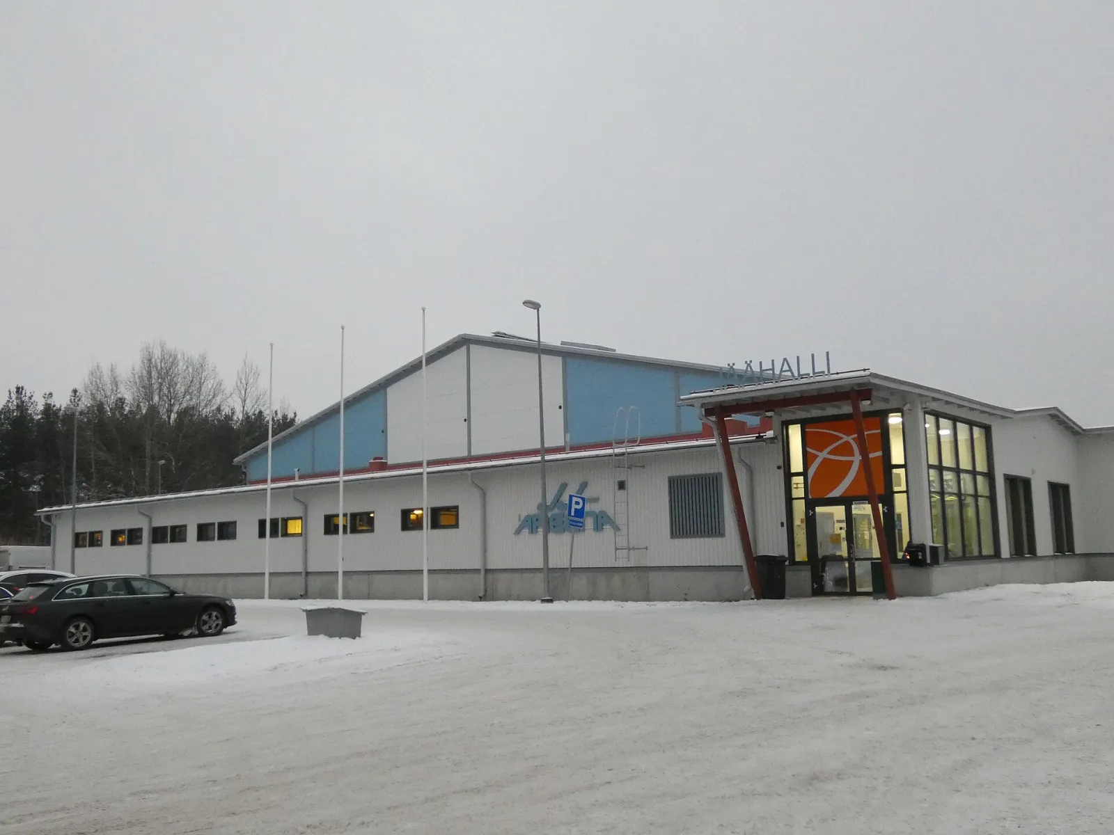 The entrance of the Riihimäki ice hall with its JÄÄHALLI sign, and the blue and grey cladding of the halls behind, seen across a snowy car park at dusk. — Riihimäen jäähalli