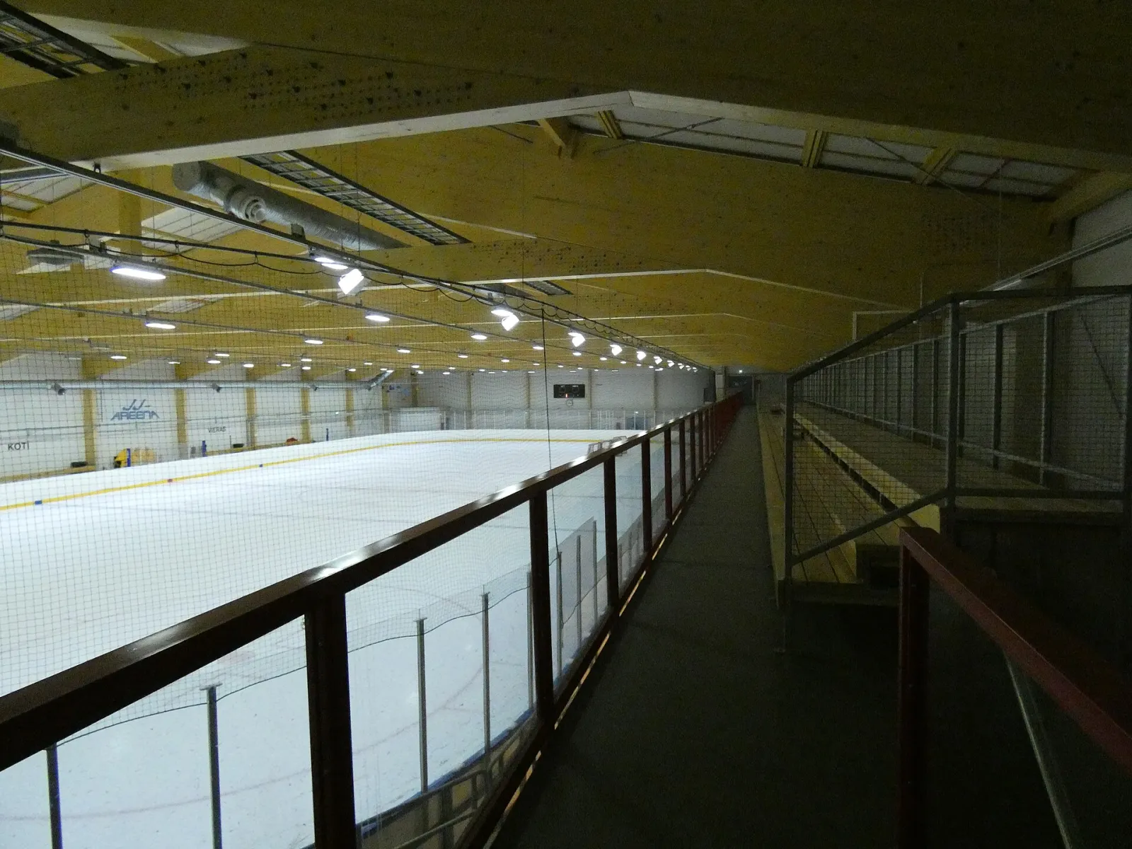 The ice sheet of the Riihimäki ice hall seen from the raised walkway along the boards, under a yellow steel roof. — Riihimäen jäähalli