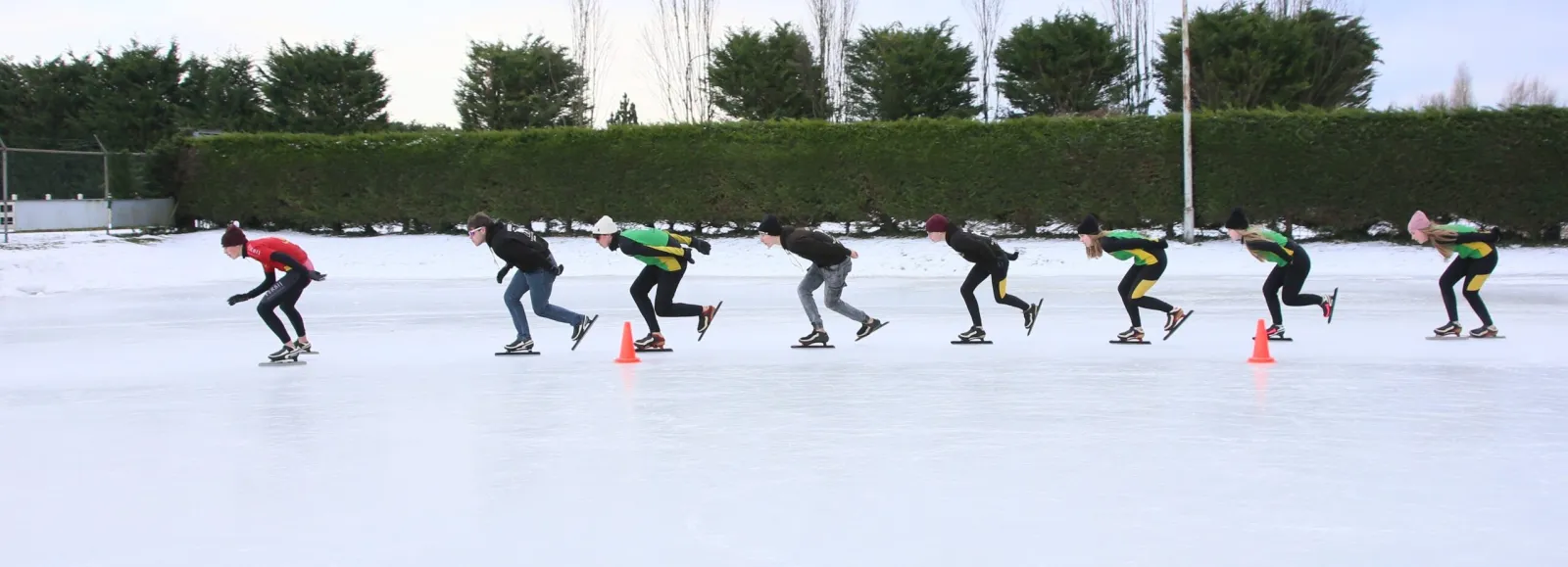 A line of skaters in club kit training in single file past orange cones on the white natural-ice rink at Rijnsburg, with a tall hedge and bare trees along the far side. — Landijsbaan 't IJswaerd (Rijnsburgse IJsclub)