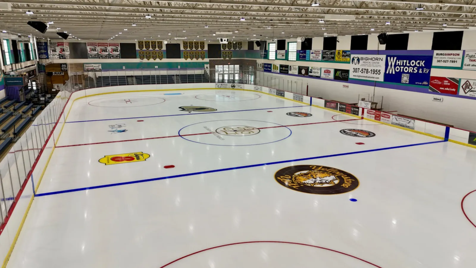 Full ice sheet from an elevated corner, blue and red rink lines and center-ice Yellowstone Quake logo visible, sponsor boards and bleacher seating along the boards, WMC.edu/cody banner readable on the wall — Riley Arena