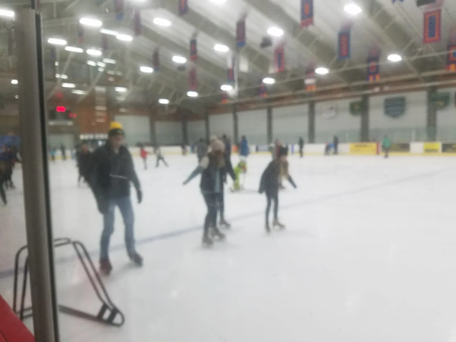 A public skate seen through the rink glass, with skaters circling under rows of hanging banners and bright ceiling lights. — Riley Rink at Hunter Park