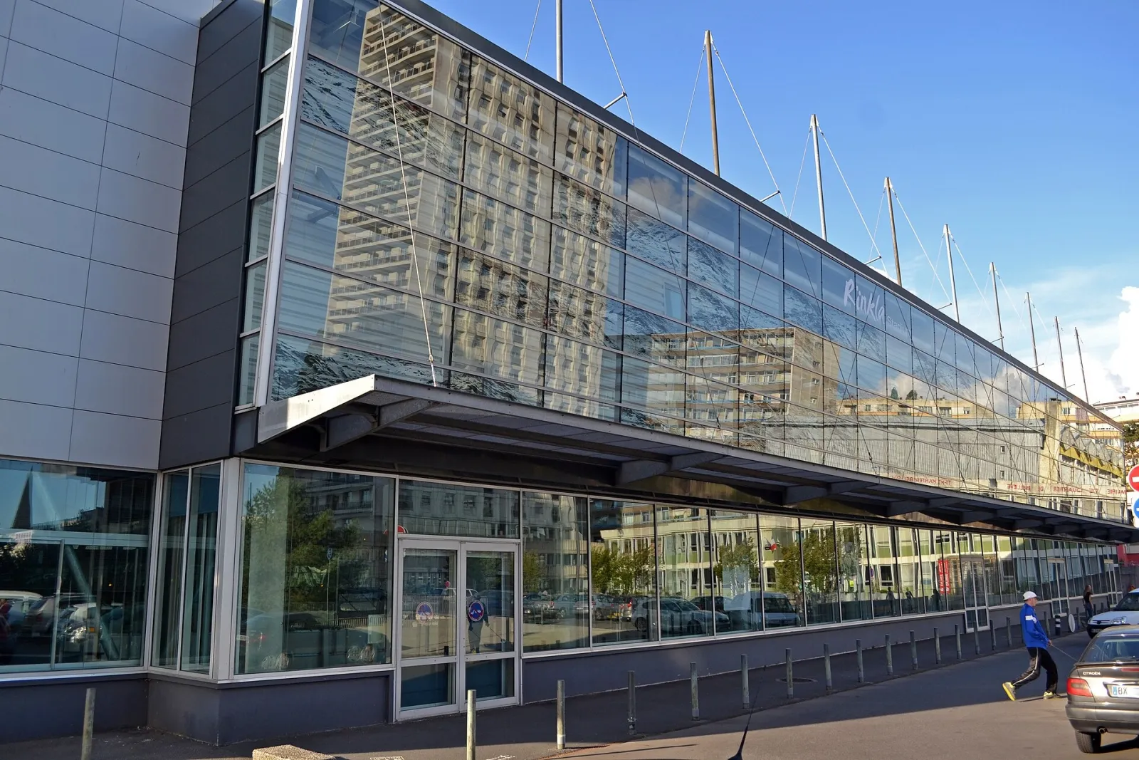 La façade vitrée du Rinkla Stadium à Brest, avec sa marquise à ossature d’acier et les câbles en forme de mât qui s’élèvent au-dessus de l’entrée. — Rïnkla