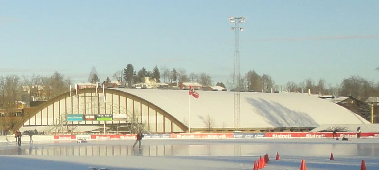 Den frosne kunstisovalen på Risenga i snøen, med hallens buede tak og flomlys langs den andre siden. — Risenga kunstisbane