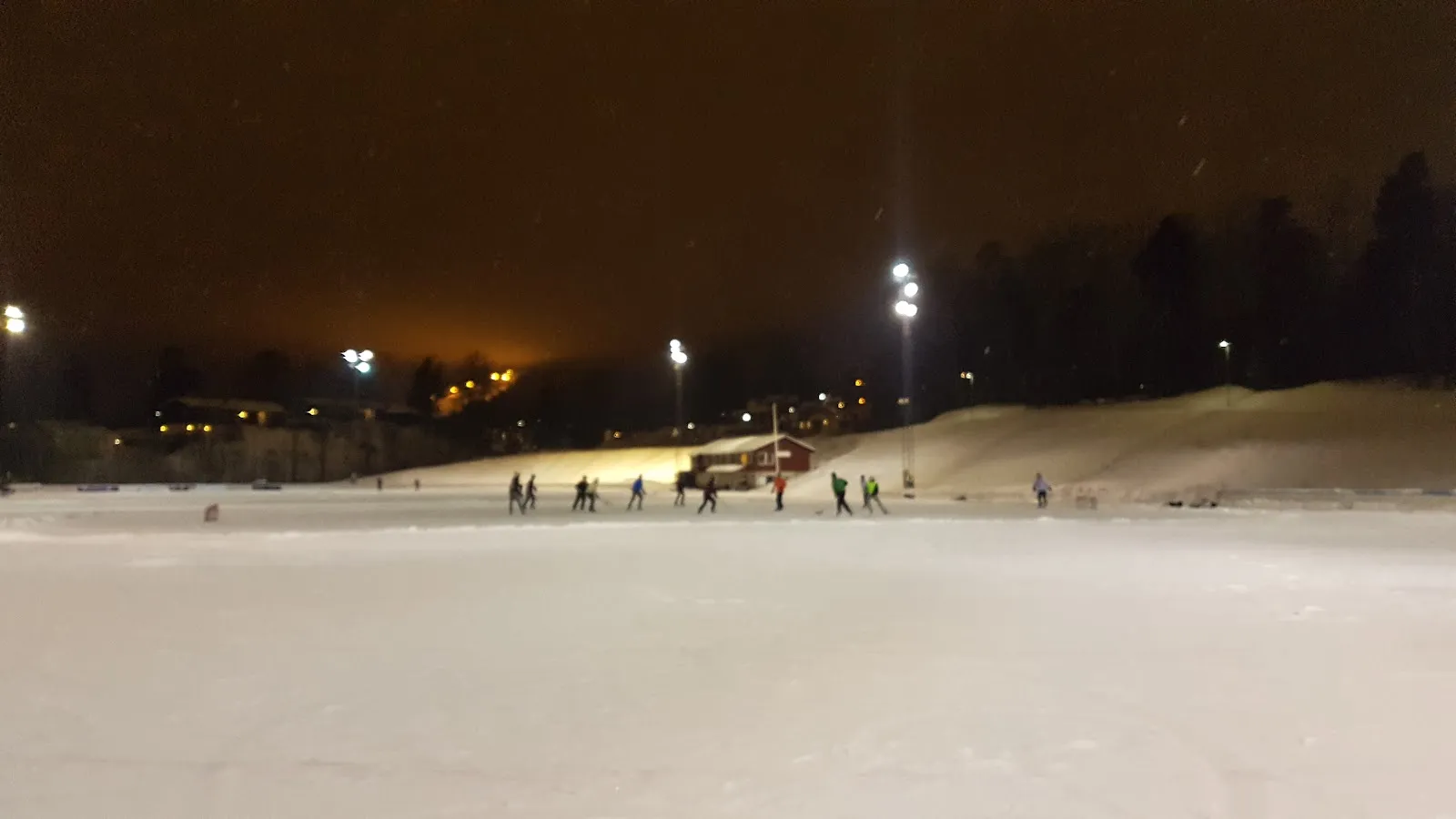 Risenga-isen under flomlys om natten, en bred, åpen bane med noen få personer på skøyter i det fjerne og åssiden bak. — Risenga kunstisbane