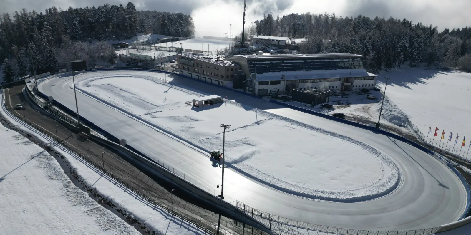 Veduta aerea dell’anello di ghiaccio di 400 metri della Ritten Arena, spolverato di neve, con gli edifici dell’arena e il bosco dietro. – Ritten Arena