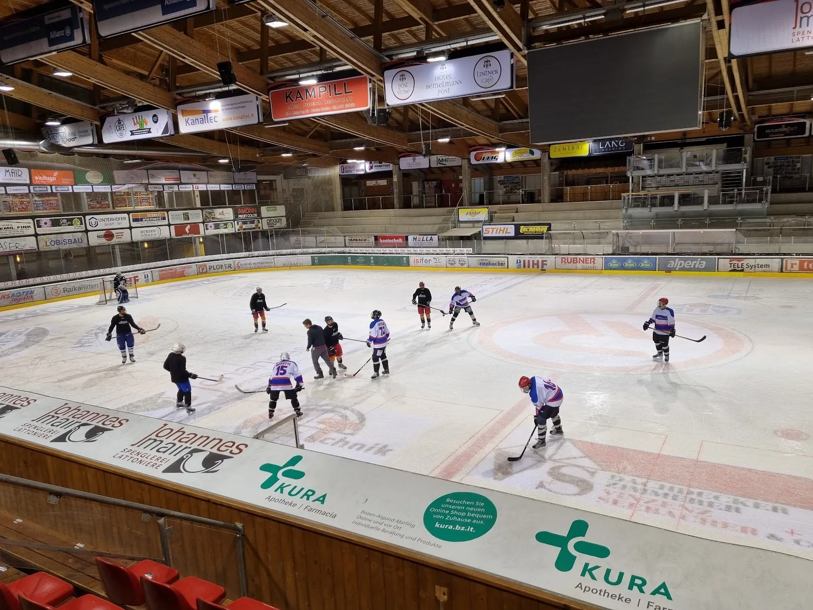Una partita sul ghiaccio coperto della Ritten Arena, con i giocatori sparsi sulla pista, i cartelloni degli sponsor e una tribuna con struttura in legno sullo sfondo. – Ritten Arena
