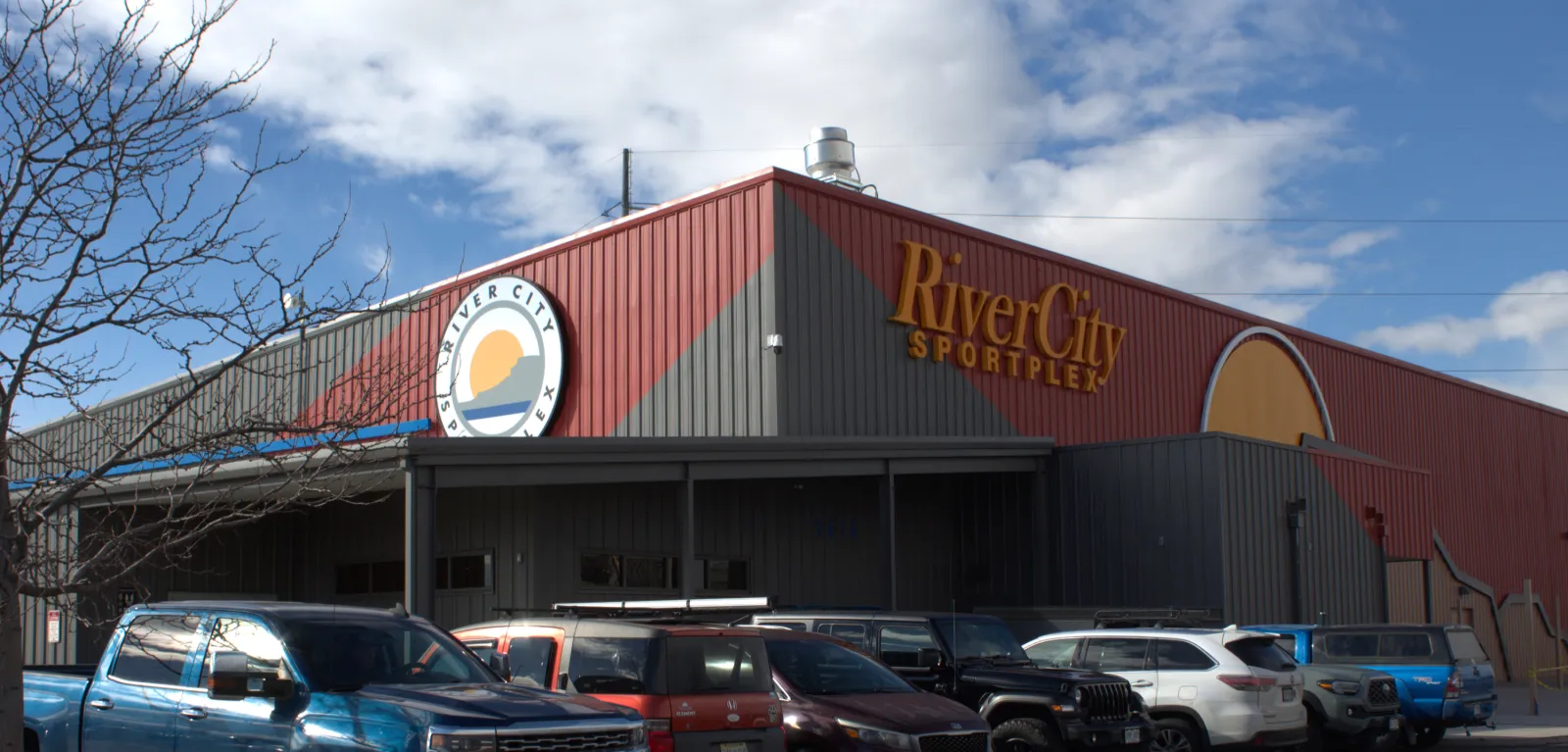 Esquina del frente del edificio de River City Sportplex, un edificio metálico rojo y gris con el logotipo circular de River City Sportplex y las letras "RiverCity SPORTPLEX" en el frontón, con vehículos estacionados al frente. — River City Sportplex