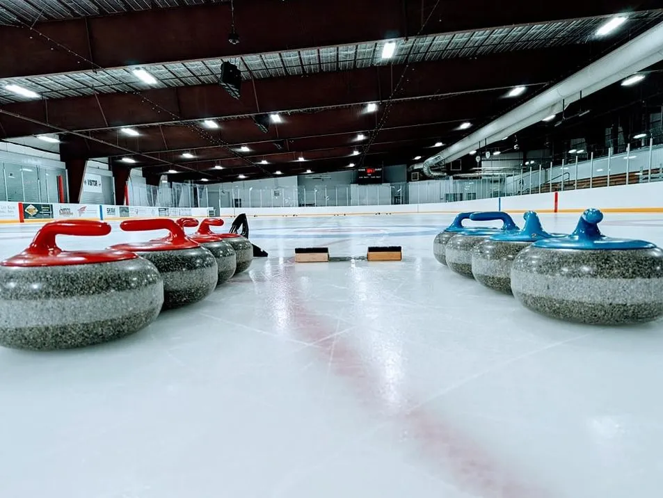 Dos filas de piedras de curling (con asas rojas y azules) colocadas sobre una pista dentro de la arena, con las vallas, gradas e iluminación superior visibles; no hay personas en la toma. — River City Sportplex