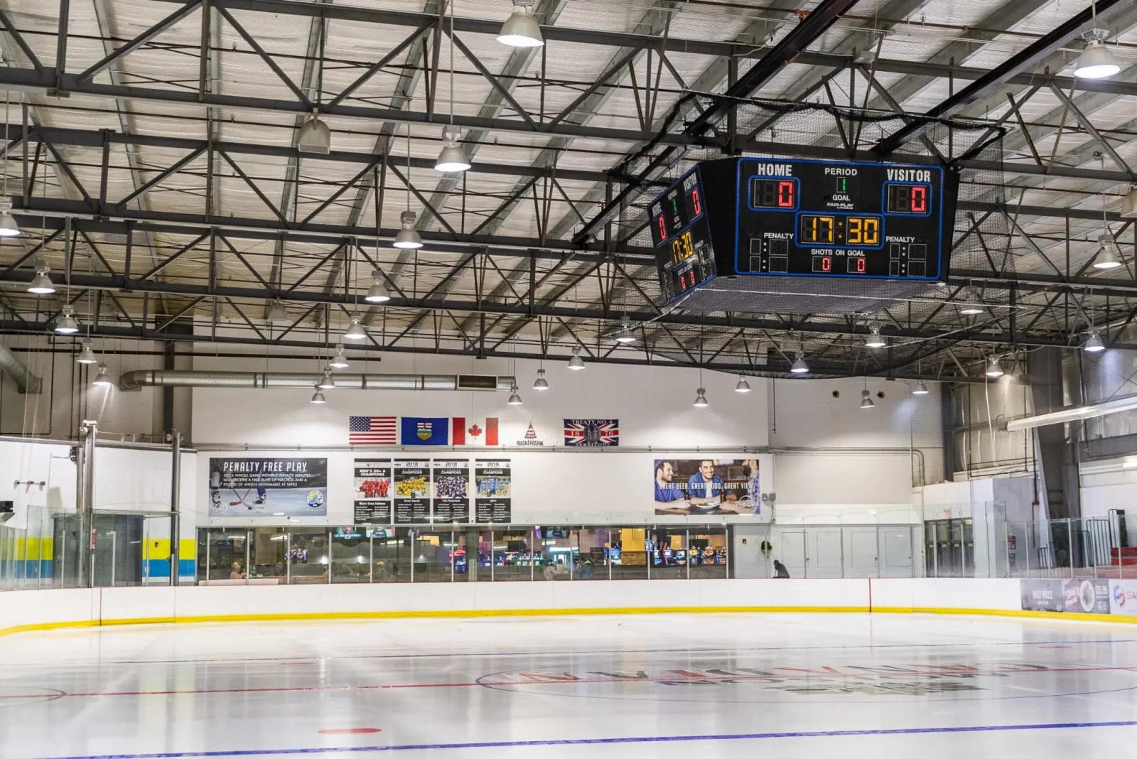 Una pista vacía en River Cree Twin Rinks, con el marcador central colgante, una fila de banderas y anuncios de patrocinadores sobre el vidrio. — River Cree Twin Rinks