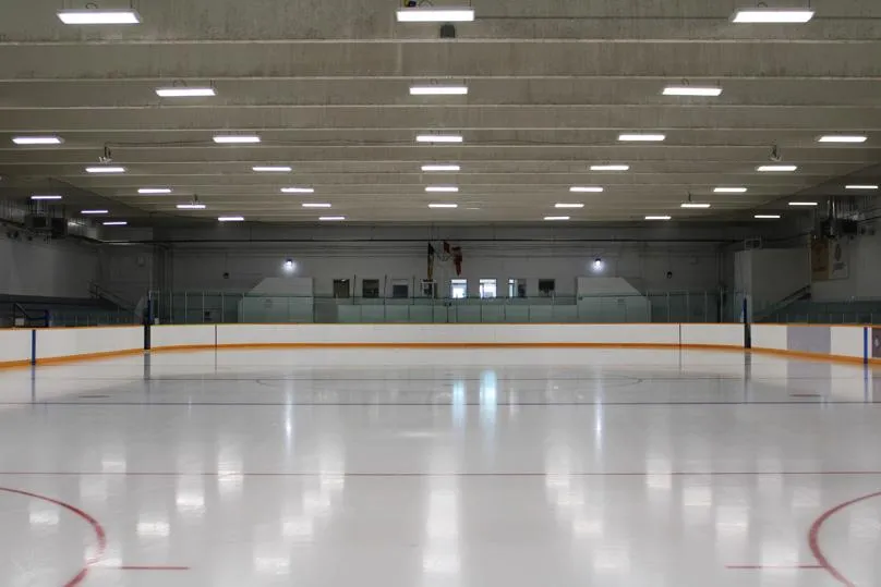 Pista de hielo vacía vista desde las gradas bajo un techo de vigas de concreto, con una bandera de Canadá y ventanas en la pared del fondo. — River East Arena