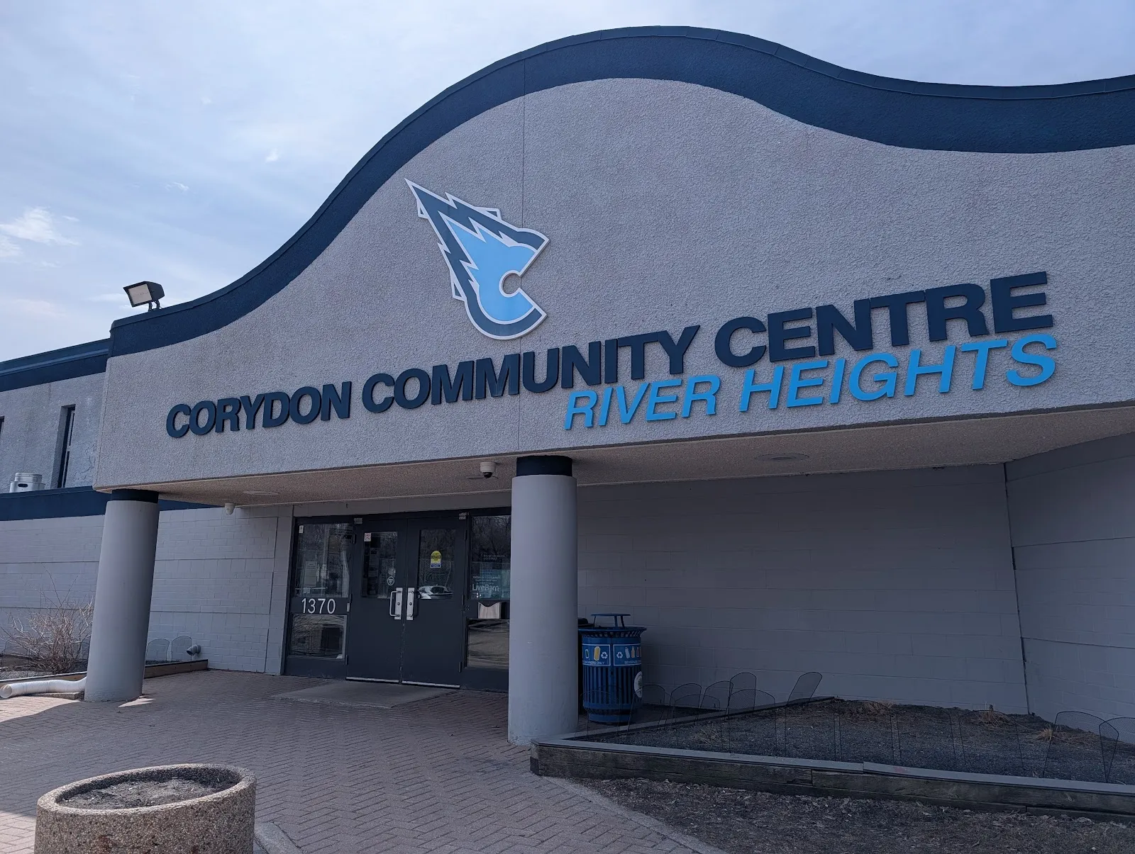 La entrada del Corydon Community Centre, sede de River Heights, en Winnipeg, con su frente curvo azul y gris y las letras del nombre — River Heights Arena (Corydon CC)