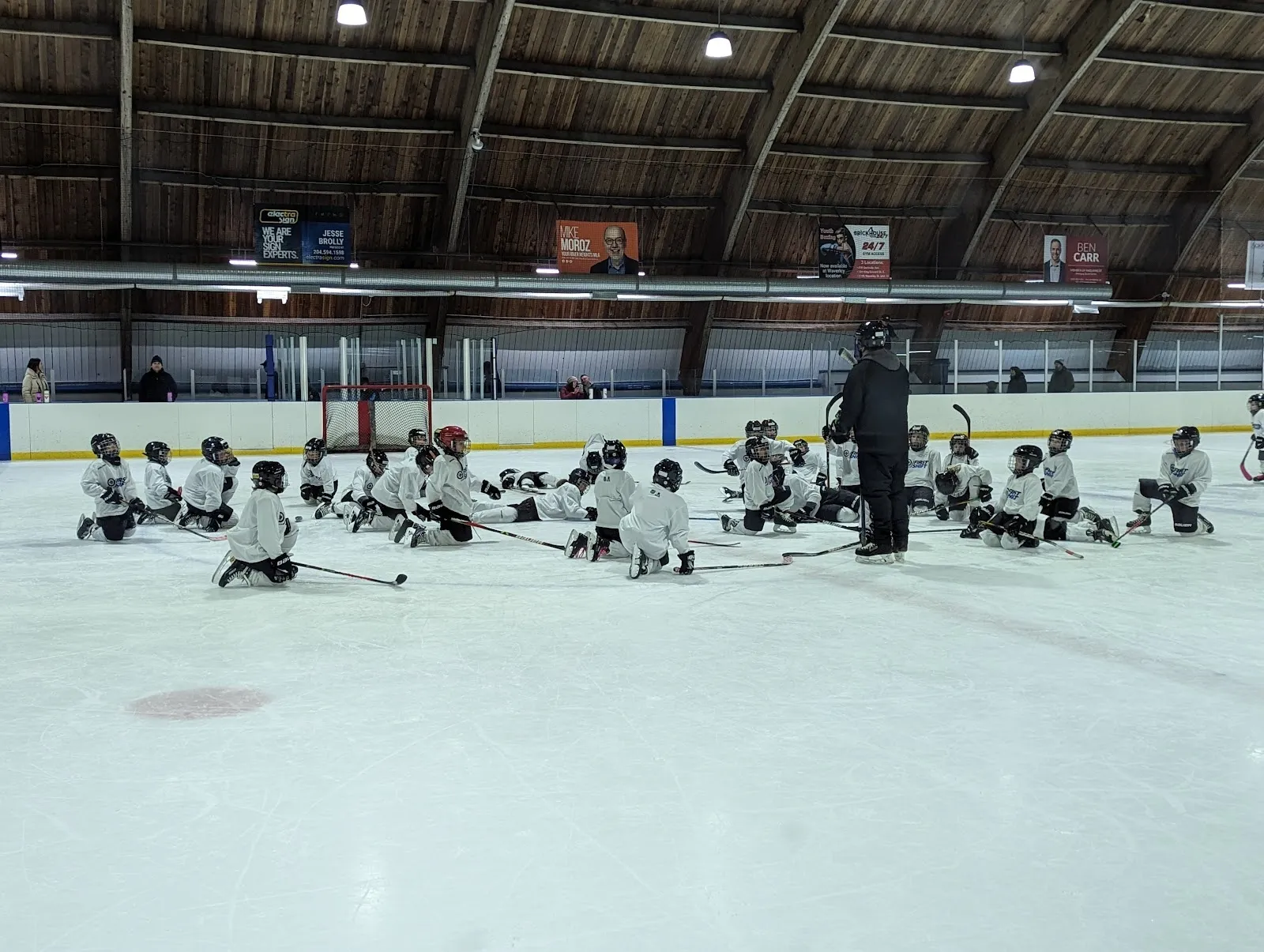 Una práctica juvenil con jugadores sentados en el hielo en hileras bajo un techo con vigas de madera, con la pista y las vallas extendiéndose detrás. — River Heights Arena (Corydon CC)