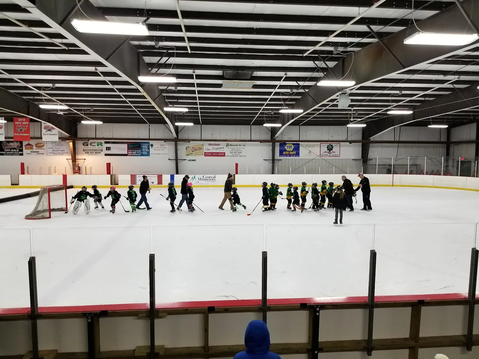 Children skating in a line along the far side of the sheet under a steel-trussed roof with sponsor boards and netting behind the goal, red boards in the foreground. — River Lakes Civic Arena