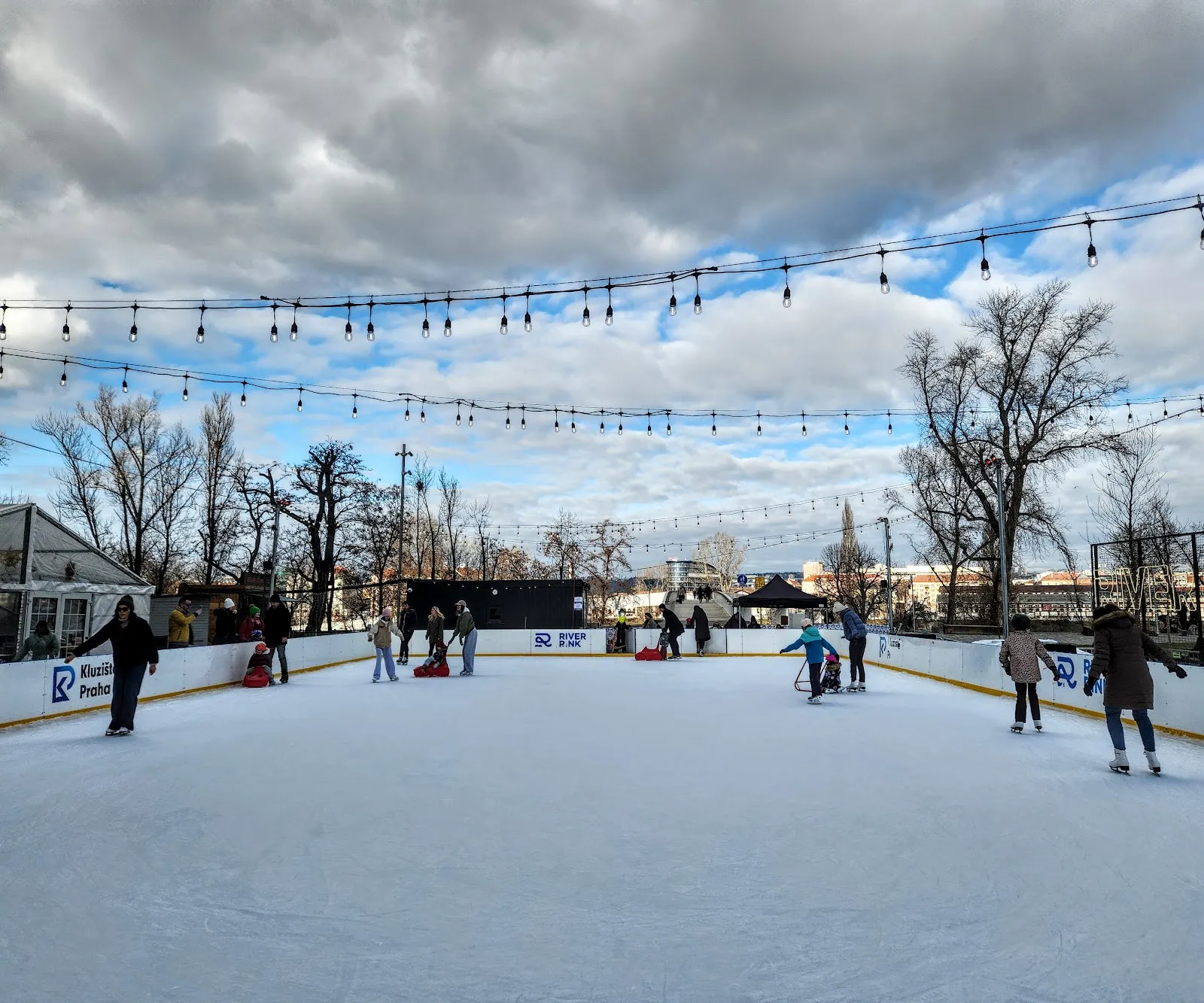 Kluziště pod širým nebem ve dne s girlandami světýlek nad mantinely, bruslaři rozptýlení po ledě a za nimi holé stromy. – River Rink Karlín