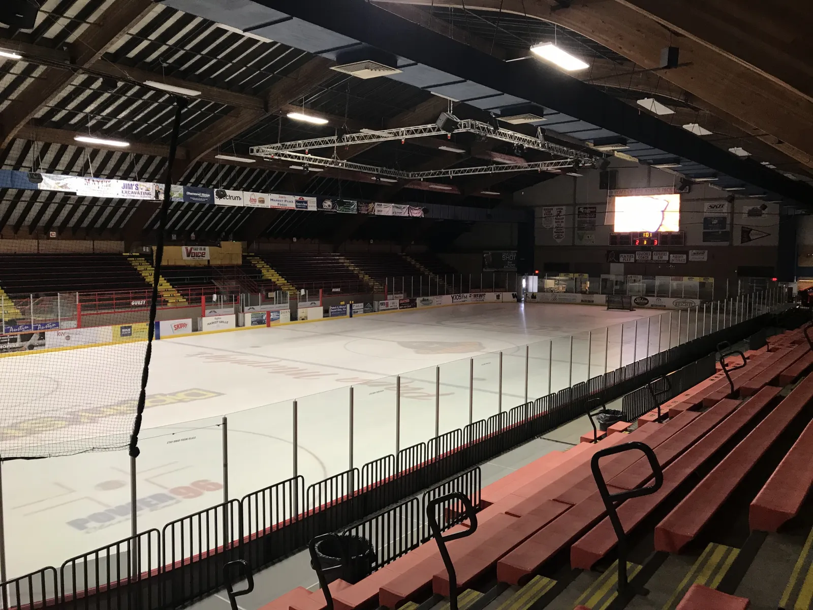 La pista vacía vista desde los asientos superiores, con hileras de gradas rojas, el cristal y un marcador bajo las armaduras de madera del techo. — Riverside Arena