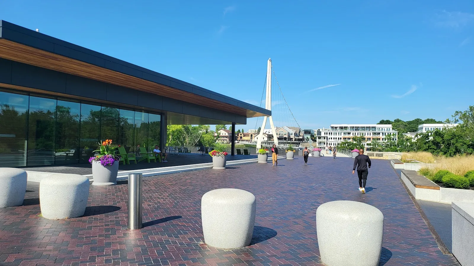 The plaza at Riverside Crossing Park in summer: a glass-walled pavilion with a timber roof on a brick-paved terrace, with the mast of the Dublin Link bridge in the distance. — Riverside Crossing Park Ice Rink