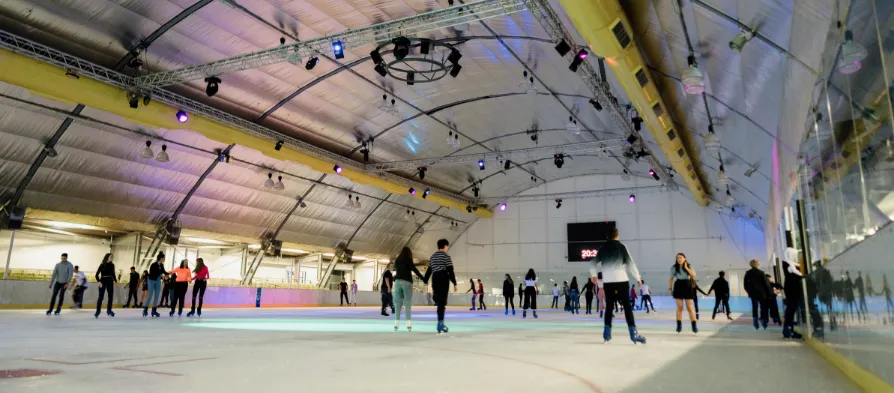 Skaters circling the ice at Riverside under a curved white ceiling with stage lighting, with the scoreboard and spectator rail beyond the boards. — Riverside Ice Rink (Riverside Leisure Centre), Chelmsford