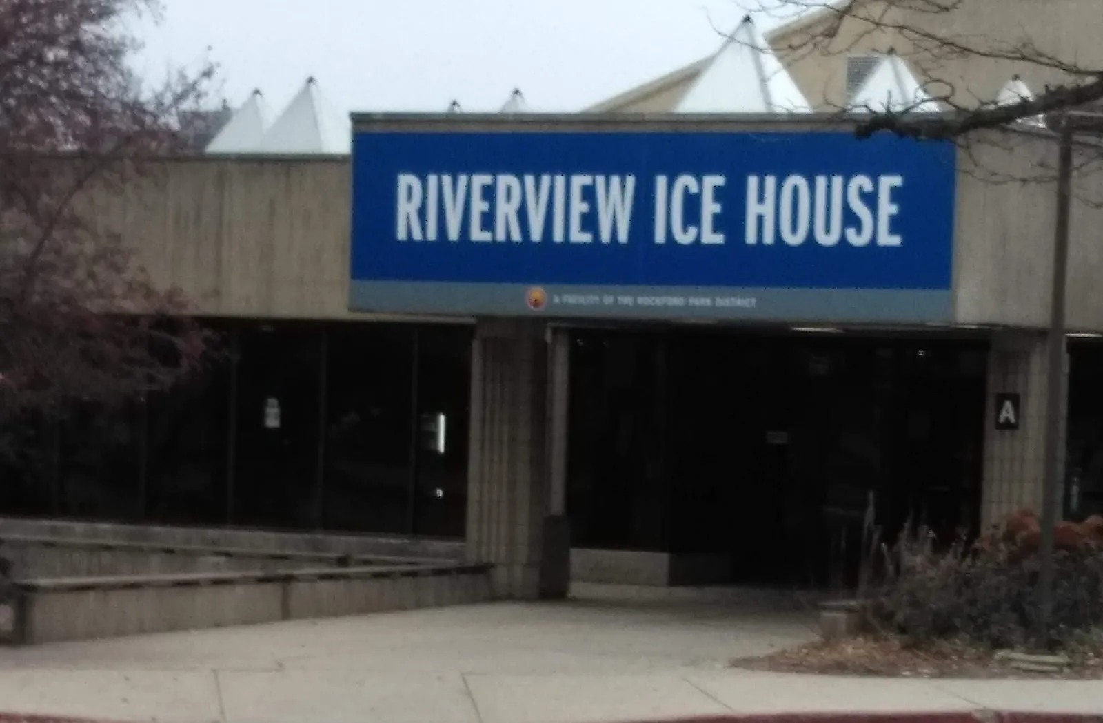 The Riverview Ice House entrance: the blue name sign over the dark glass doors of the low concrete building, with the white roof peaks behind — Riverview Ice House