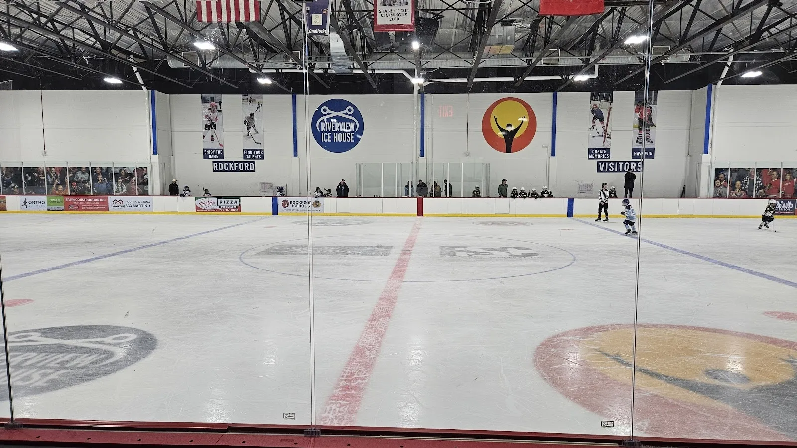 A wide empty sheet with faceoff circles and a centre red line, banners hung on the far wall below a steel-trussed roof. — Riverview Ice House