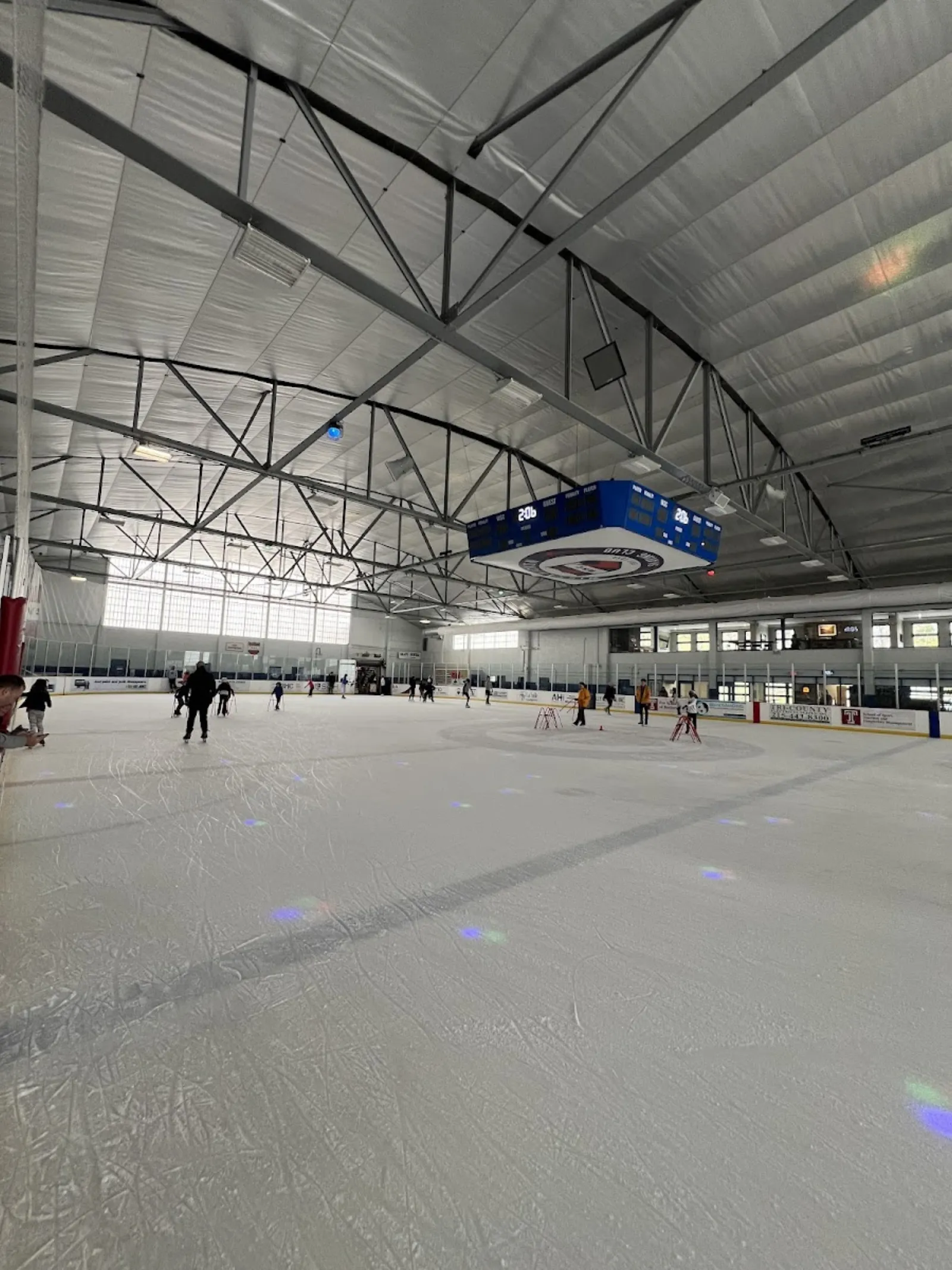 Una vista amplia de la pista desde las bandas, bajo el techo de acero en arco, con un marcador sobre el centro del hielo y algunos patinadores a lo lejos. — Rizzo Rink