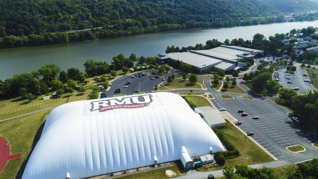 Aerial view of the white fabric-domed rink with a large 'RMU Robert Morris' logo on its roof, the attached grey-roofed main building behind it, parking lots either side, and the Ohio River and tree-lined far bank in the background. — RMU Island Sports Center