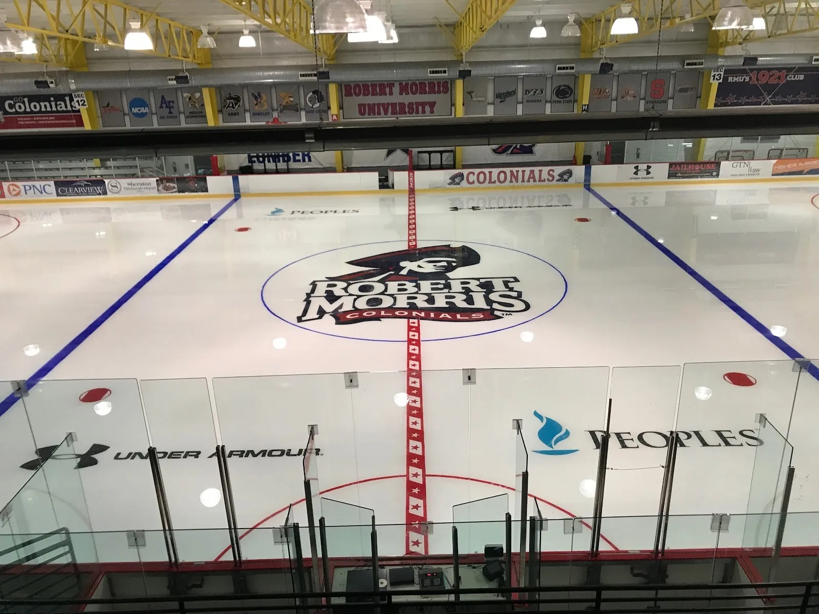 The empty sheet from behind the glass with the RMU Colonials logo at centre ice, red and blue lines and sponsor boards around the rink. — RMU Island Sports Center
