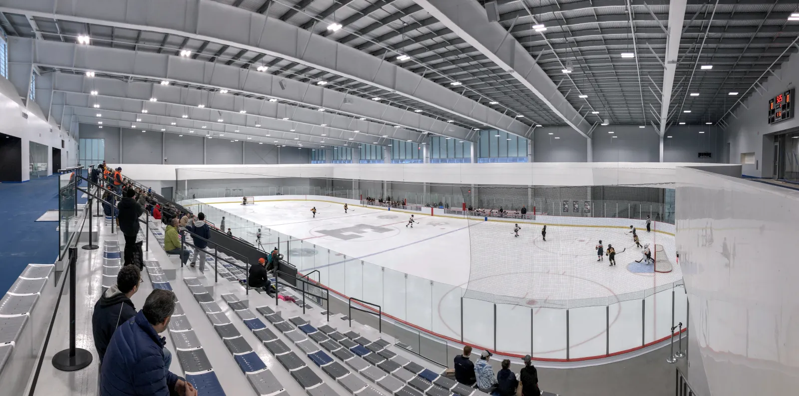 Rink 1 seen from the top of the seating bank: the whole sheet with a youth hockey game in progress, the spectator seats, the scoreboard and the netting above the boards — Robert Crown Community Center