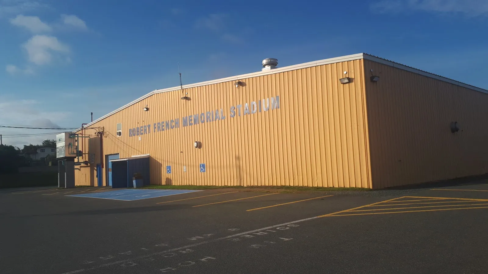 The golden-yellow metal-clad Robert French Memorial Stadium with its name lettered along the wall, in warm evening light with an empty paved lot and accessible parking in front. — Robert French Memorial Stadium
