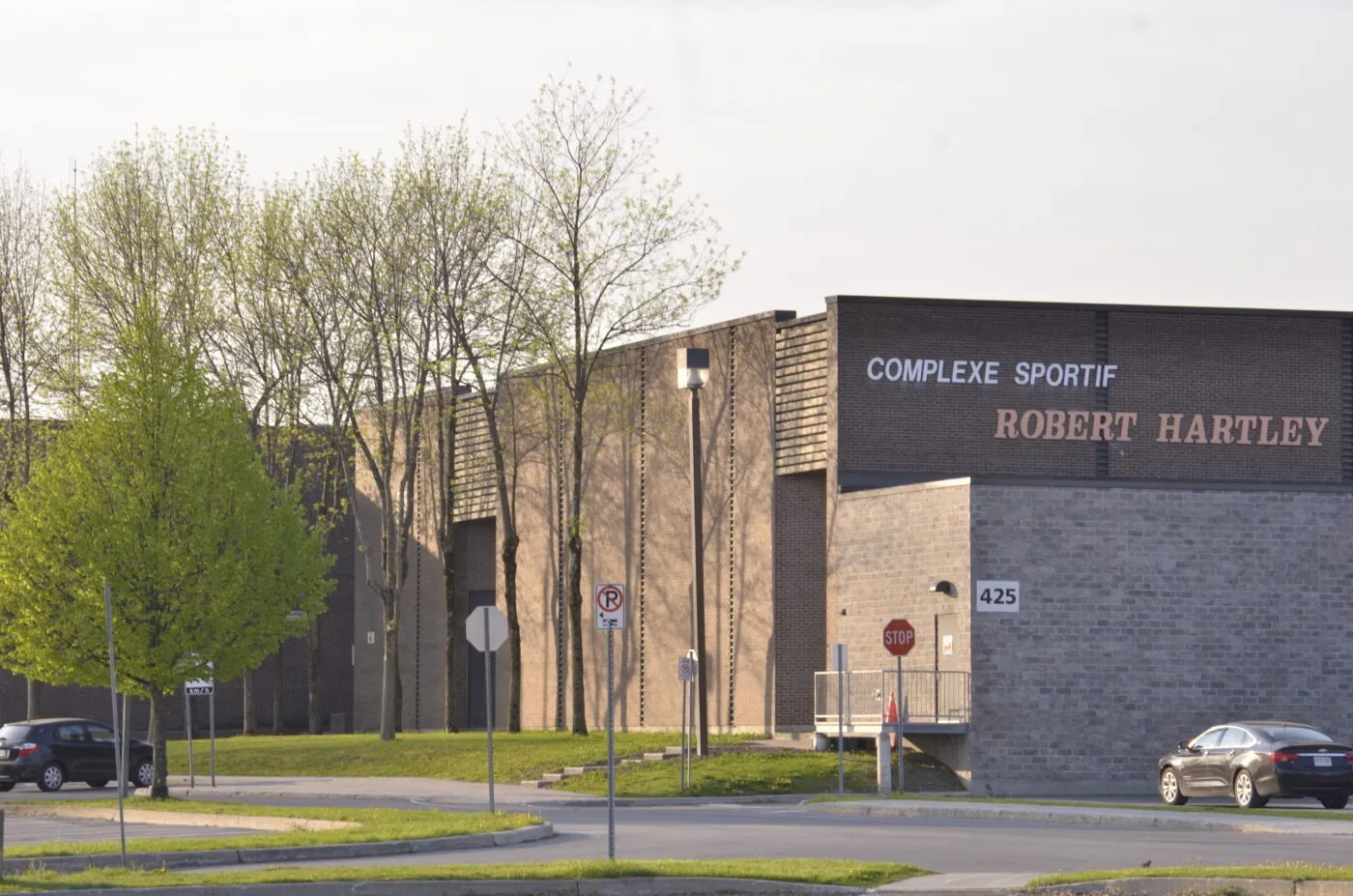 Brick and dark-block facade of Robert Hartley Sports Complex with the Complexe sportif Robert Hartley sign above a side entrance, seen across the lawn from the car park. — Robert Hartley Sports Complex (Hawkesbury)