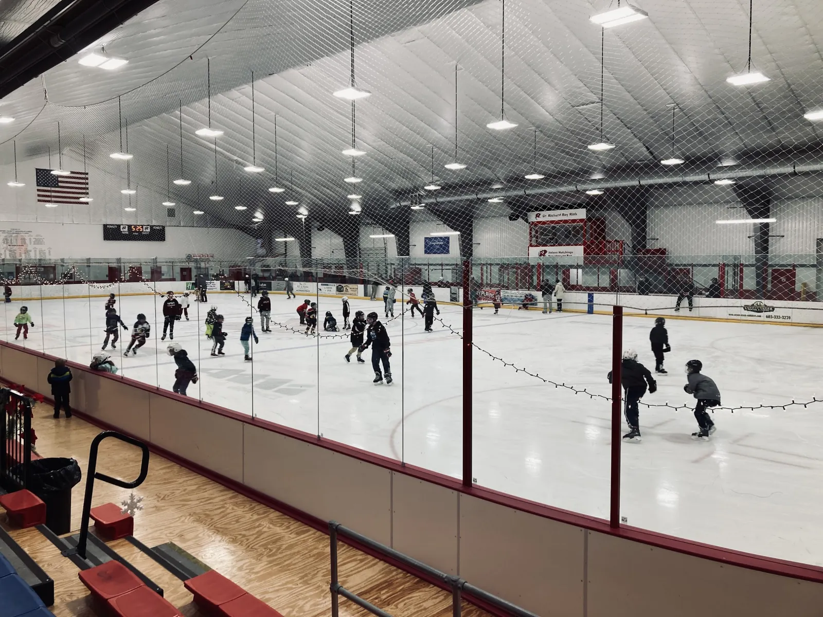 Una sesión de patinaje vista desde las gradas detrás del cristal, con una bandera de Estados Unidos y mantas en la pared del fondo bajo un techo blanco en arco. — Rochester Arena