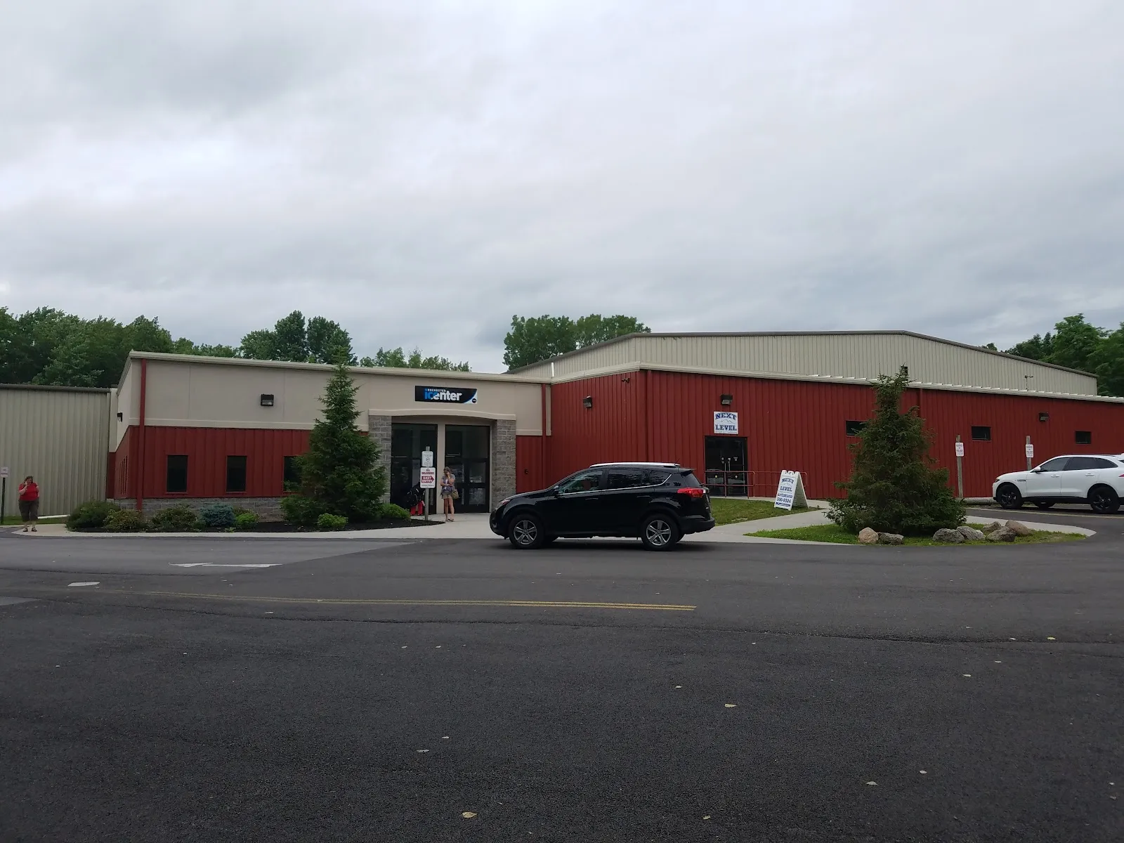 El largo edificio de Rochester Ice Center con revestimiento metálico rojo y color arena, con su entrada principal, árboles y un auto estacionado al frente, visto desde el otro lado del estacionamiento. — Rochester Ice Center