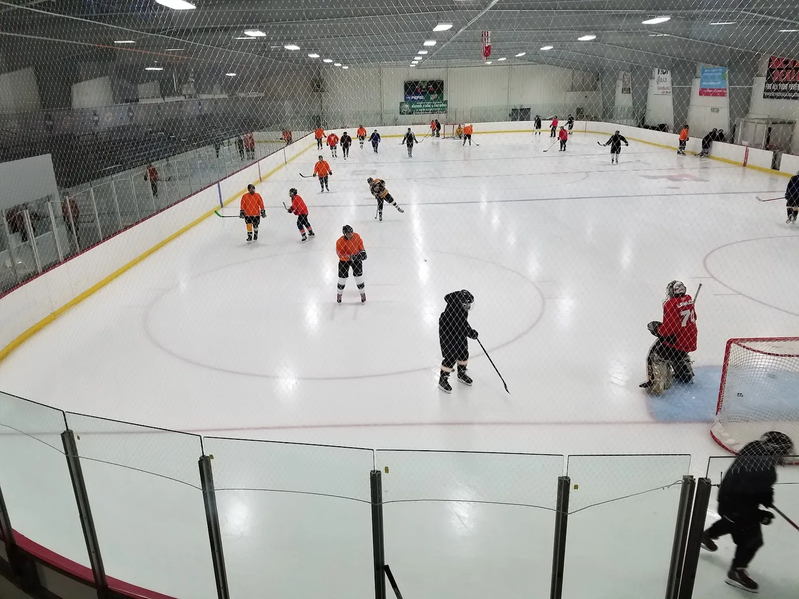 Una vista amplia de un partido visto por encima del vidrio, con jugadores repartidos por el hielo blanco bajo un techo de acero con colores de equipo rojo y naranja cerca de las vallas. — Rochester Ice Center