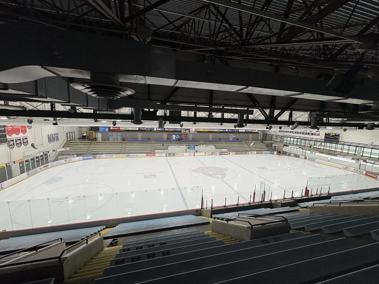The empty ice sheet inside the Rochester Recreation Center arena, with tiered seating along one side and the roof trusses above. — Rochester Recreation Center