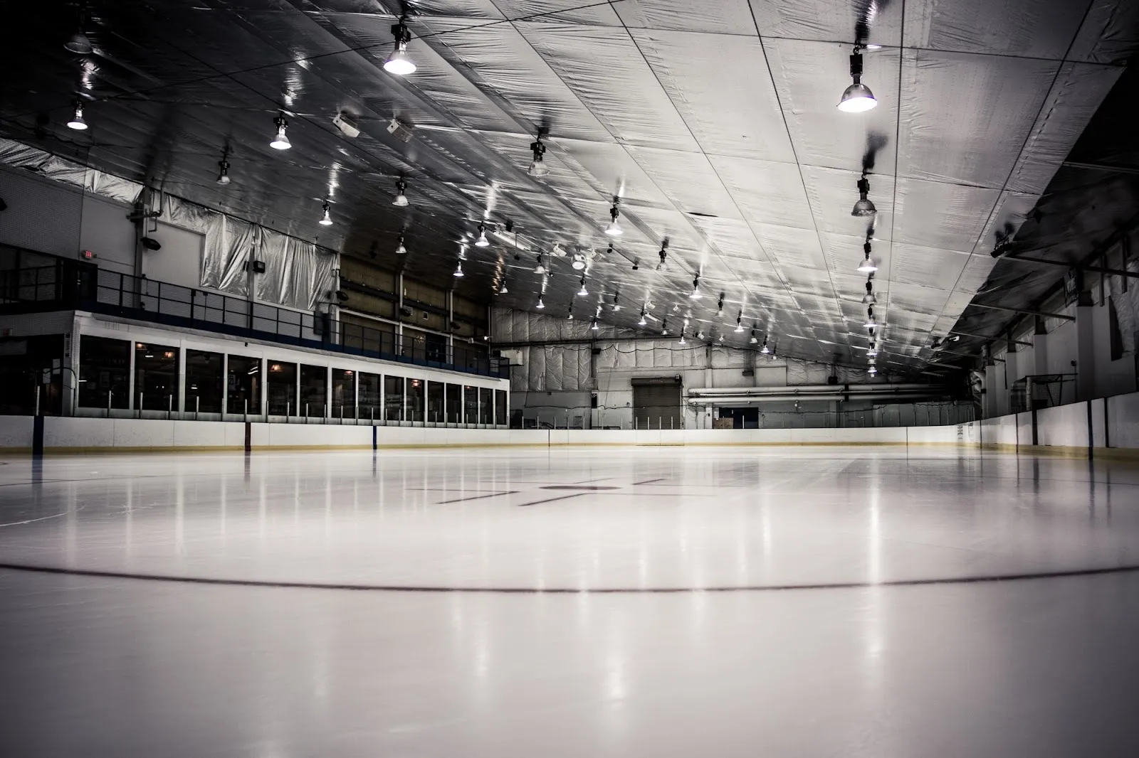 An empty sheet seen from the end, a long dark ceiling hung with lights over the white ice and boards along the left side. — Rocket Ice Skating Rink (Bolingbrook)
