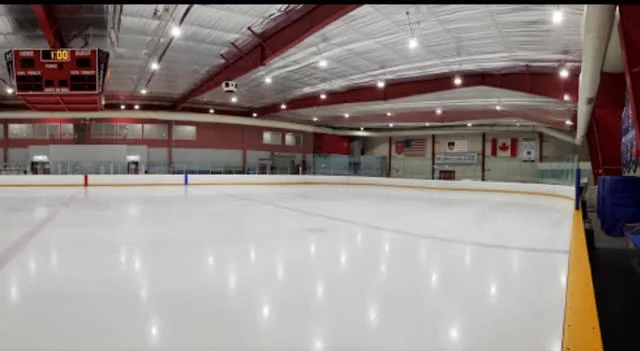 La superficie de hielo vacía de Rockville Ice Arena bajo vigas rojas del techo, con el marcador y banderas sobre las vallas. — Rockville Ice Arena