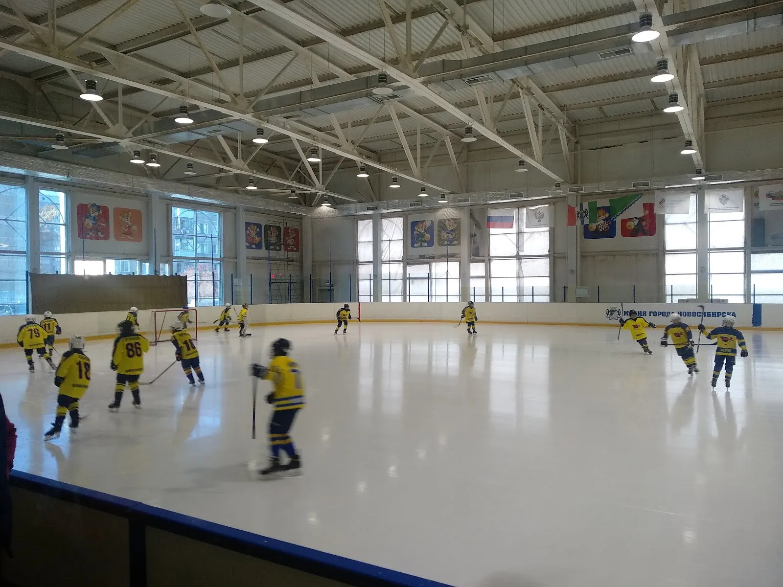 The Rodnik ice arena during a youth hockey session: the white ice sheet with boards and a long steel-truss roof, and players in yellow jerseys crossing the rink — Rodnik Ice Arena Novosibirsk