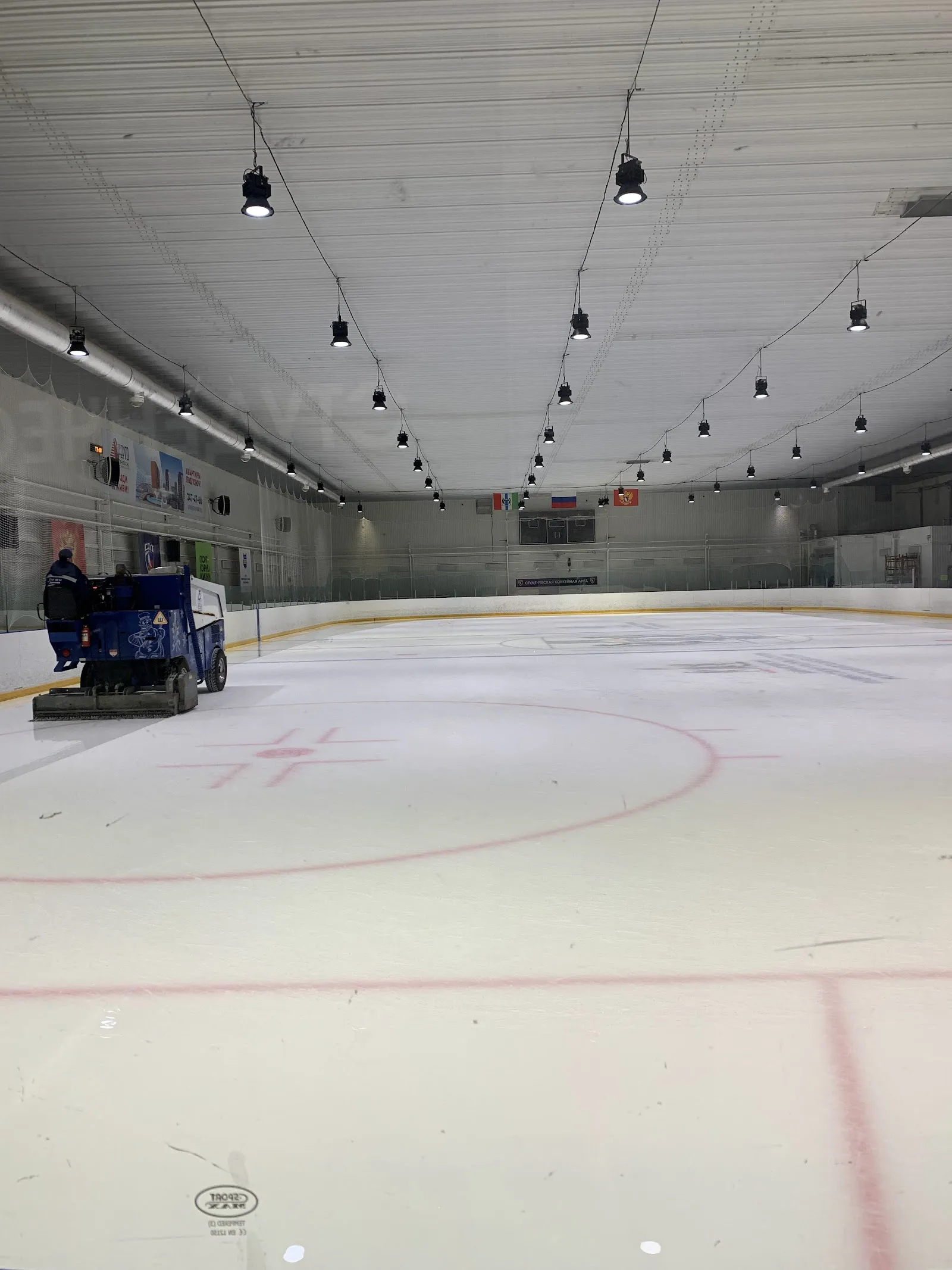 The empty ice sheet at Rodnik with an ice resurfacer parked along the boards, red goal lines and circles and rows of ceiling lights above — Rodnik Ice Arena Novosibirsk