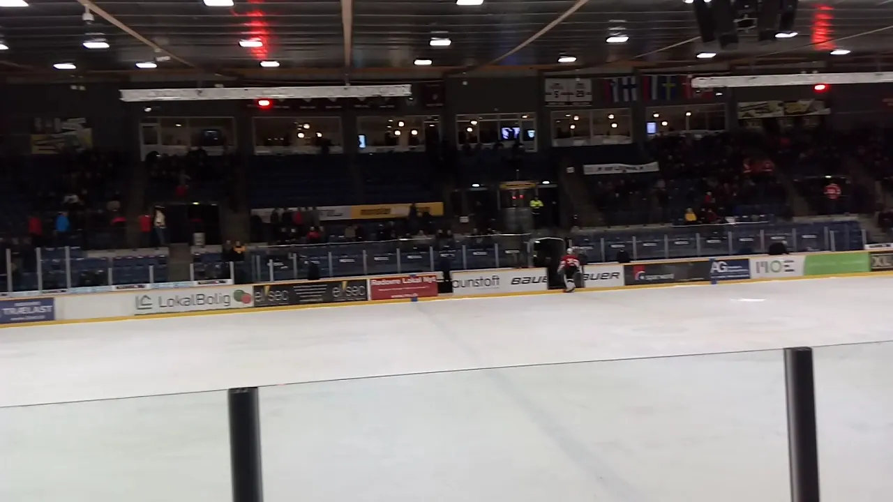 Isen i Rødovre Skøjte Arena set hen over glasset, med sponsorbander, mørke tilskuerpladser og et øvre galleri bagved. — Rødovre Skøjte Arena