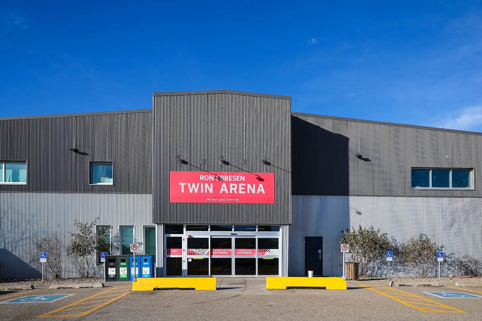 The grey cladded front of the Ron Ebbesen Twin Arena with its red Twin Arena sign above the glass entrance, seen from the parking lot under a clear sky. — Ron Ebbesen Twin Arena