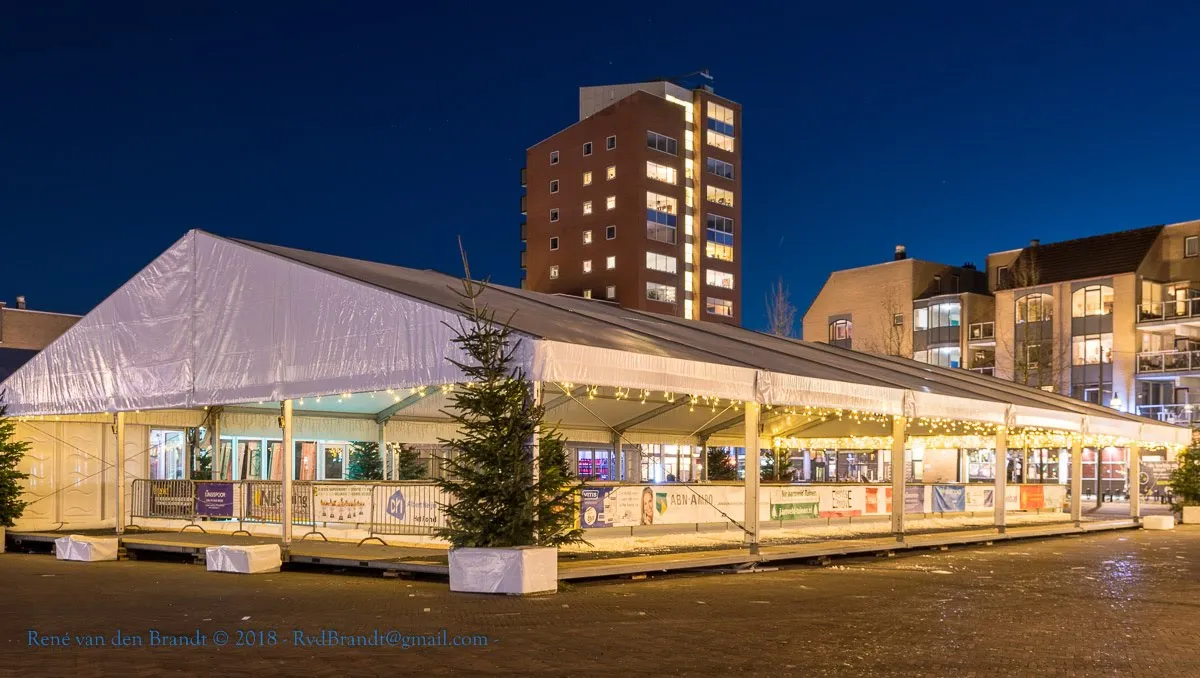 The lit white tent of Rond on Ice in the Het Rond shopping centre at night, with a Christmas tree beside it; no people. — Rond on Ice
