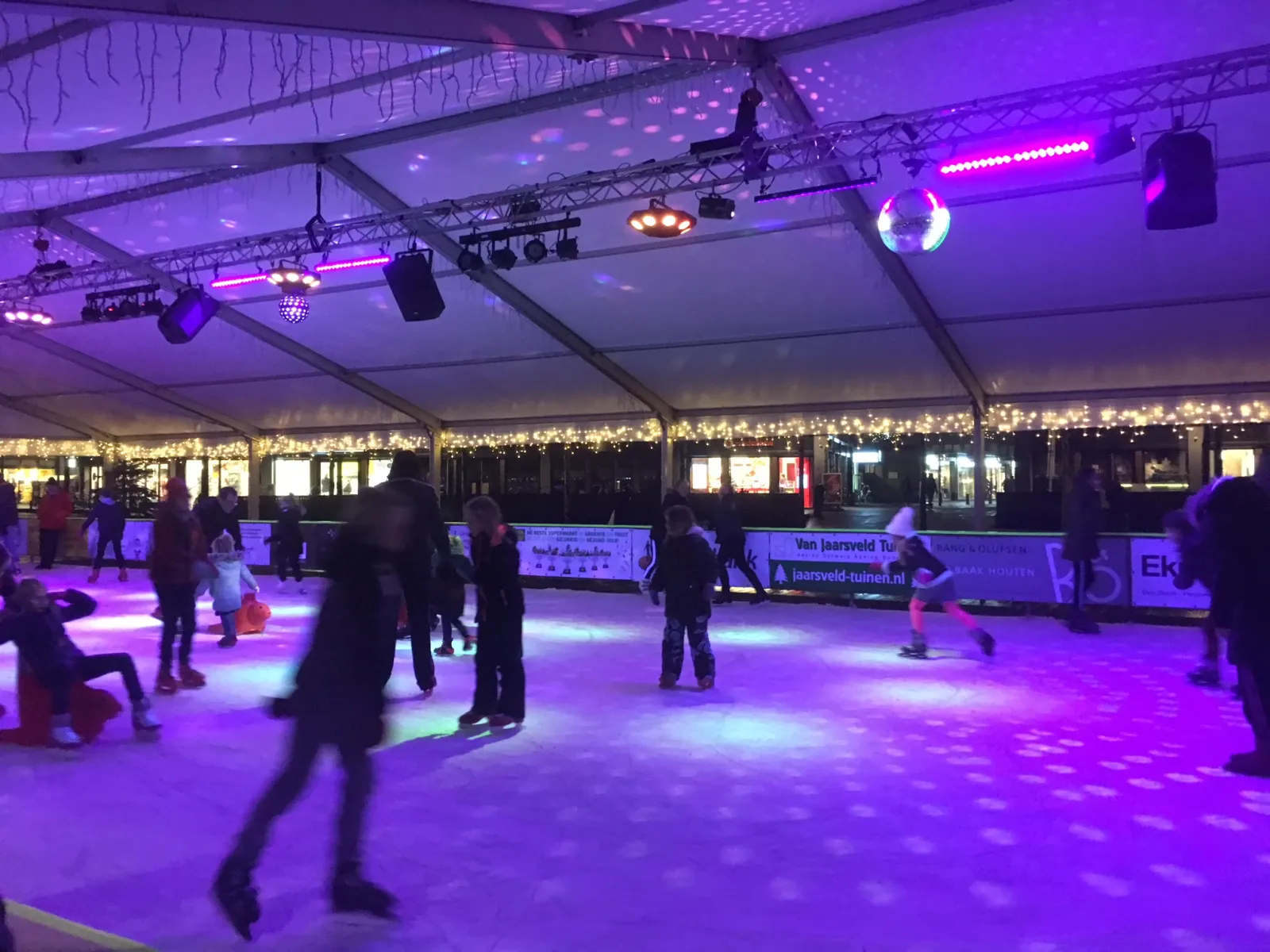 Skaters on the lit Rond on Ice rink in Houten under the tent roof; skaters at distance, not posing. — Rond on Ice