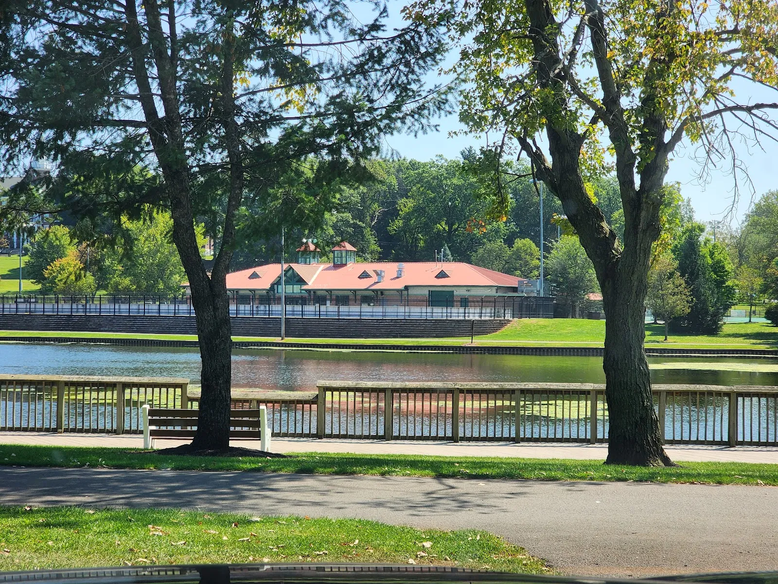 The red-roofed rink building and its fenced rink seen across the park lake, with the paved lakeside path and trees in front. — Roosevelt Park Family Skating Rink