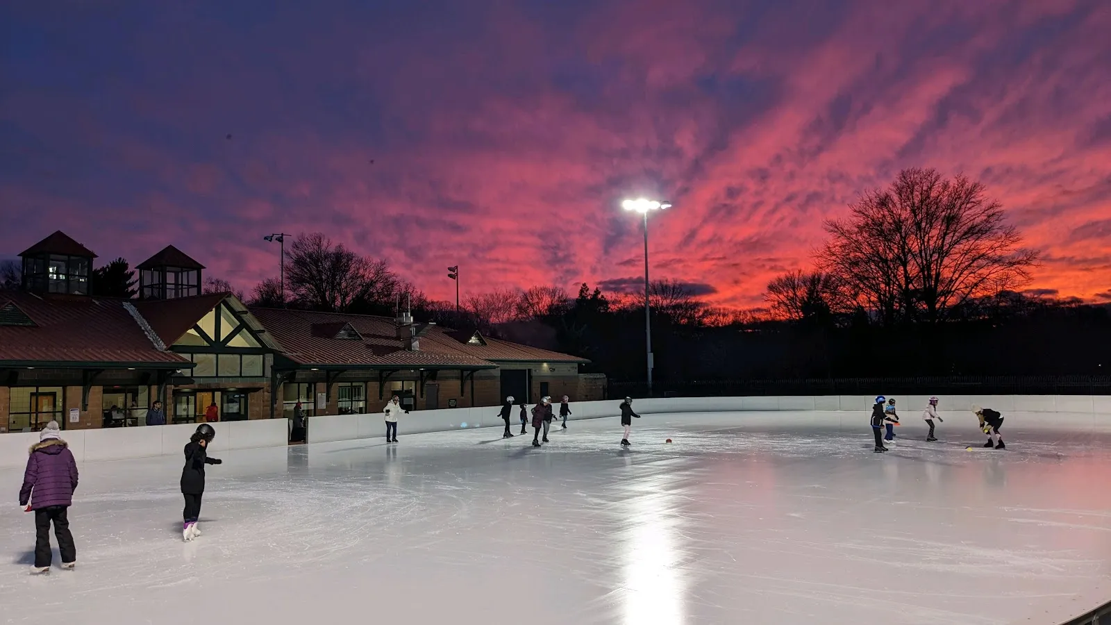 The outdoor ice at dusk under a red sky, with skaters on the rink and the timber-framed rink building and its lamp posts along the far side. — Roosevelt Park Family Skating Rink