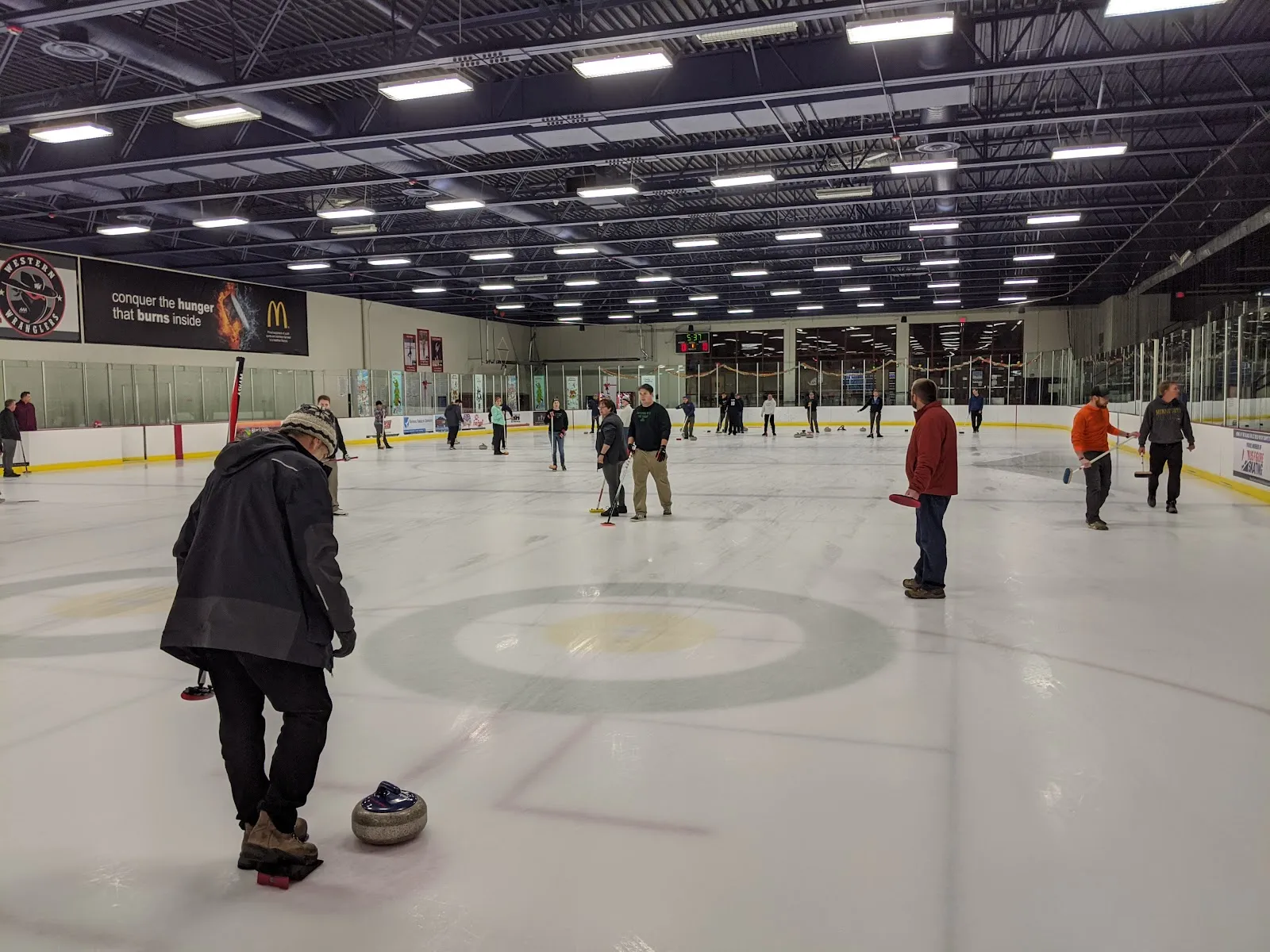 A wide view of the Roosevelt Park Ice Arena ice with the centre circle, skaters near the far boards and the arena lights above. — Roosevelt Park Ice Arena