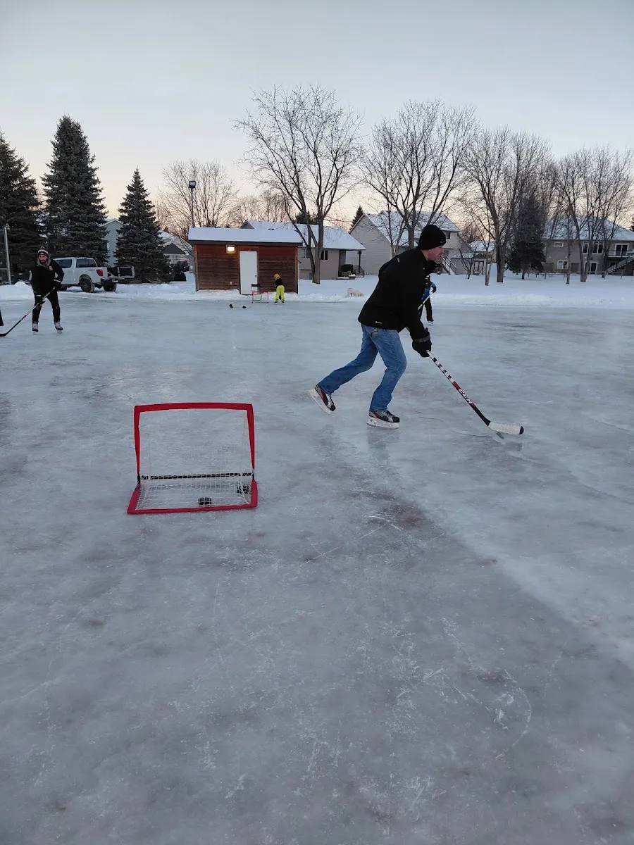A pickup hockey game on the outdoor ice at Roosevelt Park, with a portable goal on the ice, a brown warming house and houses and trees behind in winter dusk. — Roosevelt Park Ice Rink (Northfield)