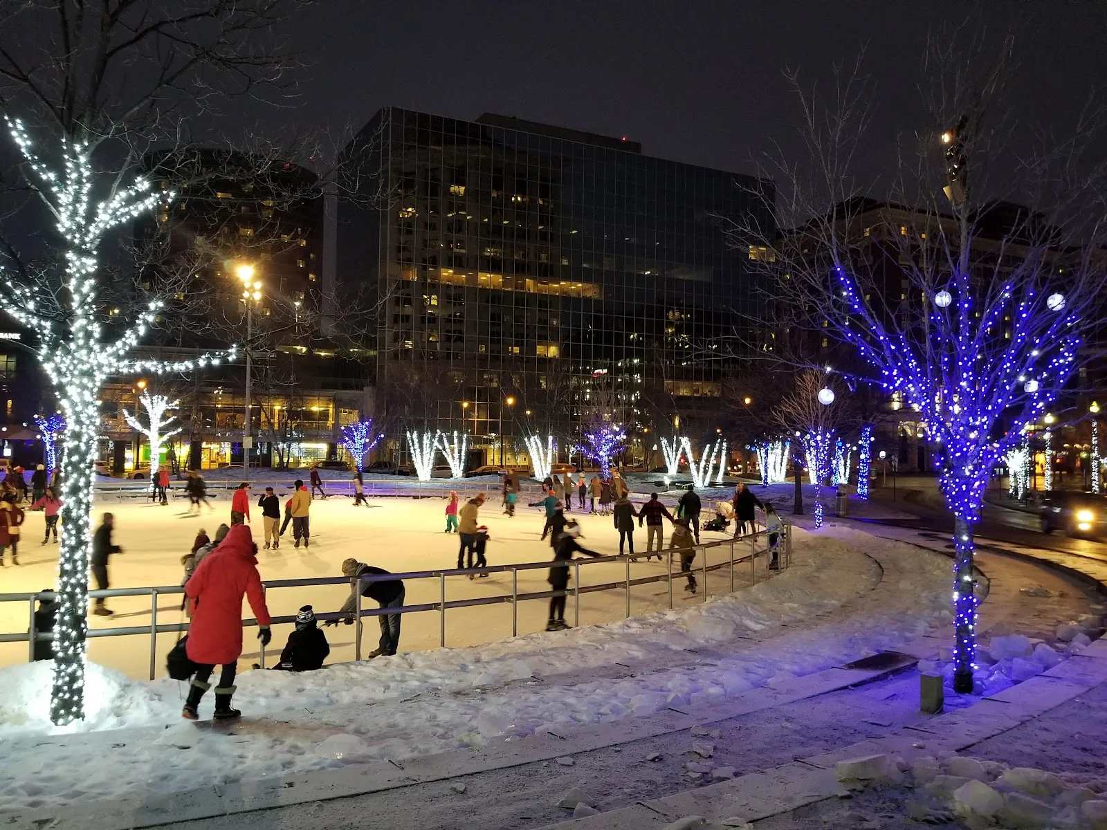The Circle's winter ice at night, ringed with lit trees and a low rail, with skaters on the ice and downtown towers behind. — Rosa Parks Circle Ice Rink