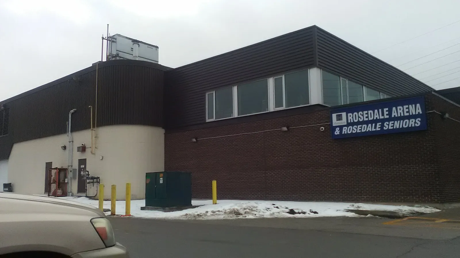 Muro de ladrillo oscuro de la arena con un letrero de "Rosedale Arena & Rosedale Seniors", bolardos y un auto estacionado al frente. — Rosedale Arena (Hamilton)