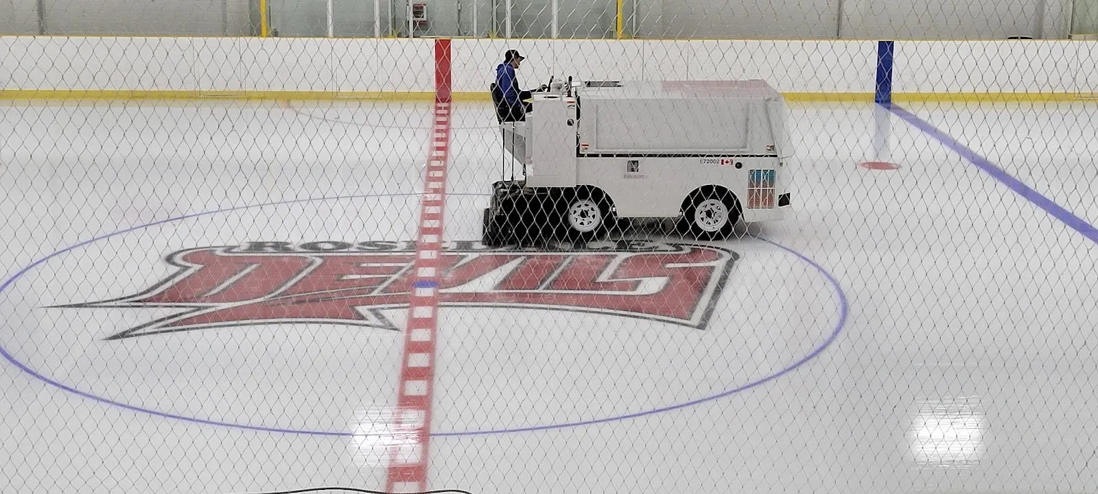 Una máquina alisadora cruzando la pista sobre un logotipo rojo de los Devils en el centro del hielo, con un techo claro y vallas blancas alrededor. — Rosedale Arena (Hamilton)